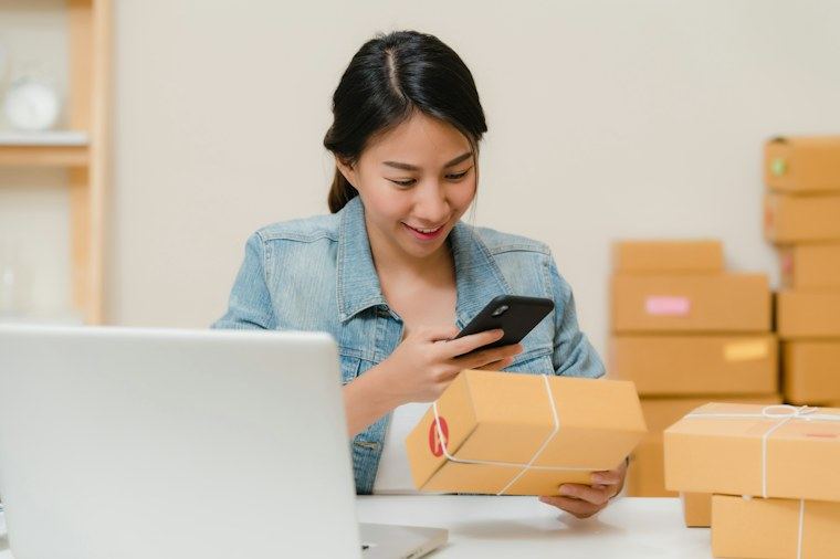 Lojista conferindo pedidos pelo celular ao lado do notebook e das encomendas